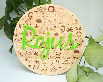 Può includere: Una targa rotonda in legno con uno sfondo marrone chiaro e un nome verde "Rojus" in corsivo. La targa è decorata con una varietà di illustrazioni di animali, tra cui giraffe, scimmie, tigri e coccodrilli.