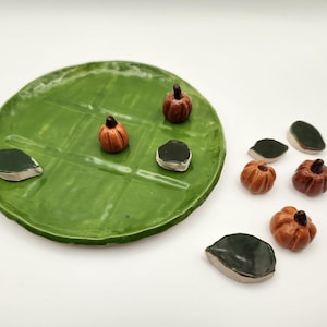 May include: A round green ceramic tic-tac-toe board with a grid pattern. The set includes two small orange pumpkins and two dark green leaf-shaped game pieces on the board, with three additional pumpkins and two leaves placed to the side.