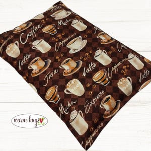May include: A brown fabric pillow with a coffee theme. The pillow is covered in a checkered pattern with coffee cups and coffee beans. The text on the pillow reads "Coffee", "Latte", "Cappuccino", "Java", "Mocha", "Espresso", and "Warm Hugs".