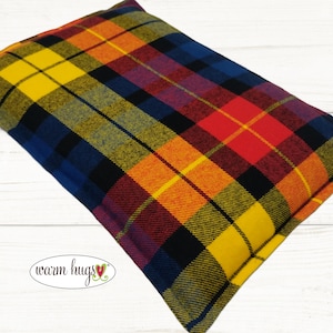 May include: A rectangular pillow with a plaid pattern in yellow, blue, red, orange, and black. The pillow has the text "warm hugs" in a cursive font with a heart symbol. The pillow is on a white wooden surface.
