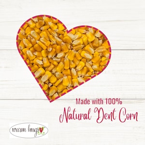 May include: A heart-shaped arrangement of yellow dent corn kernels on a white wooden background. The text "Made with 100% Natural Dent Corn" is below the heart. The text "warm hugs" is in the bottom left corner with a small red heart.