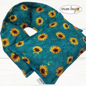May include: A teal-colored, rectangular neck wrap with a sunflower pattern. The sunflowers have yellow petals and dark brown centers. The words "warm hugs" are printed on the top right corner of the wrap.