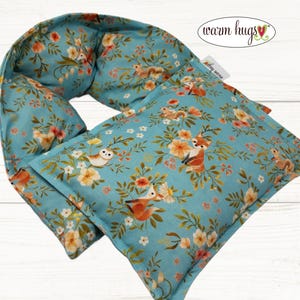 May include: A blue floral-patterned heating pad and neck wrap set. The fabric features a repeating pattern of foxes, owls, squirrels, and flowers in orange, white, and yellow. The words "warm hugs" are in a white oval.