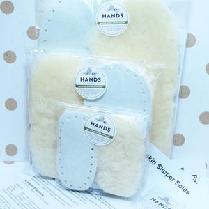 May include: Three sets of sheepskin slipper soles in different sizes, packaged in clear plastic bags. Each bag has a white label with the text "HANDS" and a website address. The text "skin Slipper Soles" is visible on a white sheet of paper.