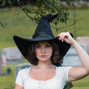 May include: A black pointed witch hat with a wide brim, a white short-sleeved top with a gathered neckline and front tie, a brown leather-look corset with lace-up detailing, and a long, solid dark green skirt worn in an outdoor setting.