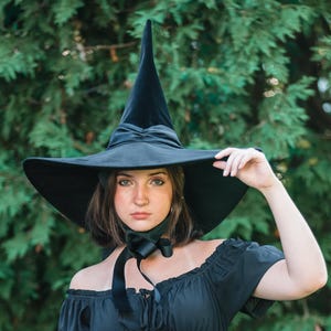 May include: A black off-the-shoulder dress featuring short puff sleeves, a fitted bodice with a central lace-up detail, and a skirt with a vertical ruffled side slit. The outfit includes a wide-brimmed black pointed hat with a ribbon chin tie.