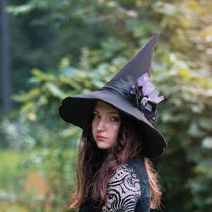 May include: A woman wearing a black witch hat with a pointed crown and a wide, wavy brim. The hat is adorned with purple accents. She is wearing a long-sleeved black dress with lace sleeves. The background is a blurred green forest.