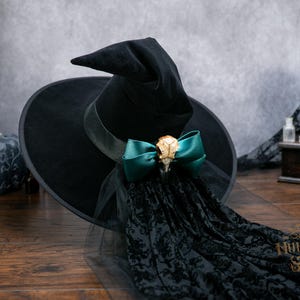 May include: A black velvet pointed hat featuring a wide brim and a tall, slightly slumped crown. A dark teal satin bow is centered on the hat band, adorned with a small, light-colored animal skull. A long, black lace veil hangs from the bow.