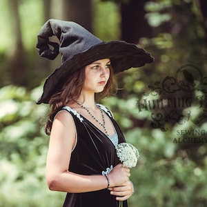 May include: A black witch's hat with a wide brim and a pointed crown. The hat is worn by a person wearing a black velvet dress with white lace trim. The image also includes the text "Hunting Faeries Sewing Alchemy".