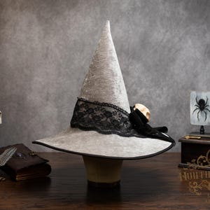 May include: A light gray witch hat with a wide brim and a black lace band. The hat is decorated with a small skull and black ribbon. The hat is part of a Halloween or gothic-themed display.