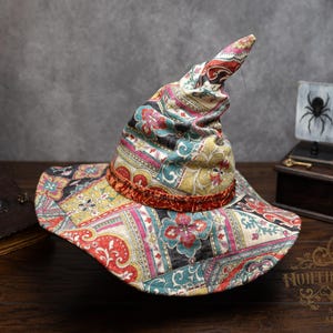 May include: A colorful witch hat with a paisley pattern in red, yellow, teal, and pink. The hat has a wide brim and a tall, pointed crown. A red velvet band accents the base of the crown. The hat is displayed on a wooden surface.