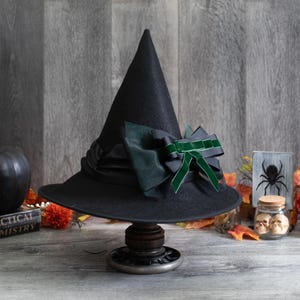 May include: A black witch hat with a wide brim and a pointed crown, adorned with a dark green and gray bow. The hat is displayed on a decorative stand, with a black pumpkin and other Halloween-themed decorations in the background.