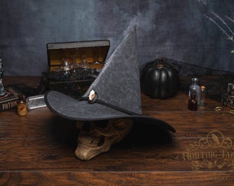 Puede incluir: Un sombrero de bruja gris con ala ancha y corona puntiaguda, apoyado sobre una calavera. El sombrero tiene un botón decorativo y un ribete negro. La escena incluye una calabaza negra, botellas de pociones y una mano decorativa.