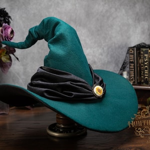 May include: A pointed teal hat with a wide brim and a slumped crown. A black fabric band is wrapped around the base of the crown, featuring a small, round, cream-colored decorative pin with a floral design centered on the front of the band.