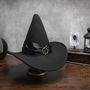 May include: A black pointed hat with a wide brim, featuring a black satin ribbon band wrapped around the base and a circular gold-toned metal buckle accent at the front center. The hat has a structured, conical crown and a slightly curved brim.