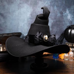 May include: A black pointed hat with a wide brim, featuring a dark velvet ribbon band tied into a bow. A small, oval-shaped brooch with a cream-colored skull motif is centered on the bow, adding a detailed accent to the solid black fabric.