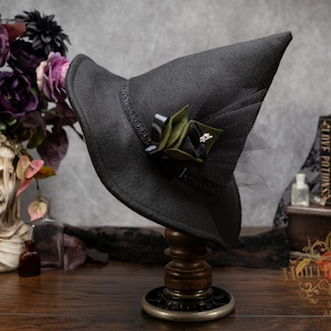 May include: A black pointed hat with a wide brim, featuring a black ribbon band, a cluster of dark green and black fabric leaves, and a small metallic accent. A layer of black netting is attached to the back of the hat, which sits on a stand.