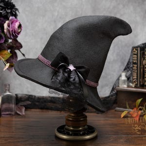 May include: A black felt witch hat featuring a pointed crown and a wide brim. The hat is decorated with a thin, textured purple band, a black velvet bow, a small silver-toned star charm, and a layer of black lace trim around the base.