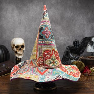 May include: A pointed witch hat featuring a vibrant, multi-colored patchwork pattern in shades of red, teal, yellow, and black. The hat has a wide, slightly wavy brim and a tall, conical crown, accented with a thin, metallic-toned cord trim at the base.