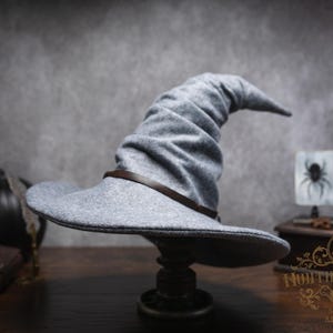 May include: A gray witch hat with a brown leather band, displayed on a decorative stand. The hat has a wide brim and a tall, pointed crown. The background includes Halloween-themed decorations like a skull, a pumpkin, and a spider.