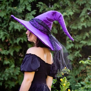 May include: A bright purple pointed hat featuring a wide brim and a tall, slightly drooping crown. The base of the crown is wrapped in a textured black lace ribbon that extends into a long, flowing black tulle veil trailing down the side.