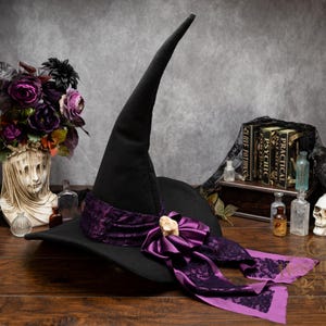 May include: A black pointed hat featuring a wide purple fabric band with a dark floral pattern and a central light-colored stone accent. The hat has a long, curved crown and a wide brim, with long fabric tails extending from the decorative bow.