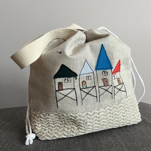 May include: A beige linen drawstring bag with a white strap. The bag features an embroidered design of four houses with different colored roofs: blue, green, white, and red.