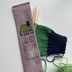 May include: A light purple fabric needle case with a house design embroidered on the front. The case has two metal eyelets and is filled with wooden knitting needles. A green and blue knitted sock and balls of green yarn are in the background.