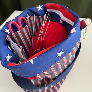 Op de afbeelding: Een breiprojecttas met een patriottisch thema, open en gevuld met benodigdheden. De tas heeft blauwe stof met witte sterren en rood-wit gestreepte stof. Binnenin zitten rode wol, breinaalden en een roze schaar.