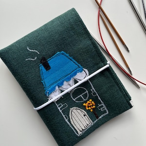 Könnte beinhalten: Eine grüne Filznadeltasche mit einem weißen gestickten Hausmotiv. Das Haus hat ein blaues Dach, eine weiße Tür und einen kleinen Baum mit orangefarbenen Blättern.