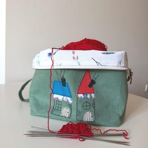 May include: A green fabric knitting bag with a floral patterned top and two embroidered houses. Red yarn spills out, with knitting needles and a small knitted piece in front. The bag has a zipper closure and a small handle.