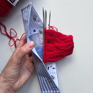 Può includere: Un astuccio per ferri da maglia a righe blu e bianche contiene diversi ferri e una calza rossa parzialmente lavorata a maglia. L'astuccio è aperto, rivelando un interno a fantasia con bottoni a pressione. Un gomitolo di filo rosso è visibile sullo sfondo.