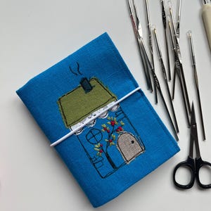May include: A blue fabric crochet hook case with an embroidered house design, secured with a white elastic band. Several silver crochet hooks and a pair of black scissors are arranged nearby, suggesting a crafting theme.