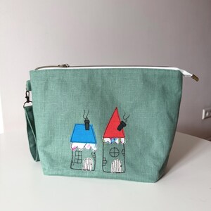 May include: A teal green zippered pouch with a white zipper pull and a small handle. The pouch features embroidered images of two whimsical houses with blue and red roofs, windows, and chimneys.