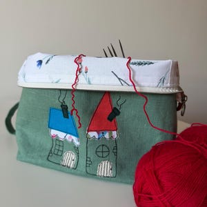 Op de afbeelding: Een groene stoffen breinaaldenetui met twee geborduurde huisjes, één met een blauw dak en de andere met een rood dak. De bovenklep is wit met een bloemenpatroon. Rood garen en breinaalden zijn zichtbaar.