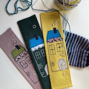 Puede incluir: Tres estuches de agujas de tela en tonos malva, verde y amarillo, cada uno bordado con un diseño de casa caprichoso. Las casas tienen tejados azules, ventanas y puertas. También se ve un ovillo de lana y agujas de tejer.