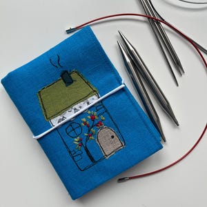 May include: A blue fabric knitting needle case with an embroidered house design, secured with a white elastic band. Silver knitting needles and red cables are also visible, suggesting a knitting project.