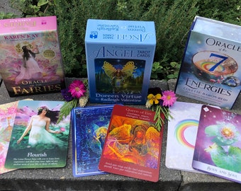 May include: A collection of oracle and tarot card decks featuring colorful illustrations of fairies, angels, and energies. The decks are arranged on a stone surface with flowers scattered around them. The decks include "Oracle of the Fairies", "Angel Tarot Cards", and "Oracle of the 7 Energies".