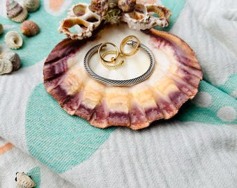 May include: Two gold rings and a silver bangle bracelet are displayed on a seashell. The seashell is on a white and turquoise patterned fabric.