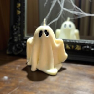 Peut inclure: Une bougie en forme de fantôme de couleur crème avec des yeux ovales noirs et une mèche. La bougie est devant un miroir, avec son reflet visible. Décoration d'Halloween. La bougie est sur une surface en bois, avec un miroir encadré noir en arrière-plan.