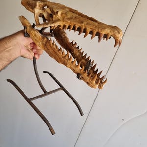 Op de afbeelding: Een gedetailleerde, bruinachtige replica van een dinosaurus schedel, met scherpe tanden en een verweerde uitstraling. De schedel is gemonteerd op een donkere metalen standaard, wat een opvallend displaystuk creëert. De open mond onthult de ingewikkelde details van de gefossiliseerde structuur.
