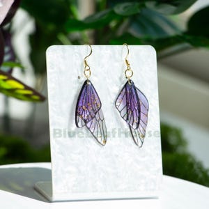 May include: A pair of drop earrings featuring a butterfly wing design. The wings are purple and translucent with gold accents and black outlines. The earrings hang from gold-coloured hooks. The text "Blue Wolf House" is visible on the display stand.