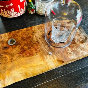 Può includere: Un vassoio di legno con bordo naturale, con una campana di vetro e un bicchiere decorativo. Il vassoio presenta un motivo a venature naturali con un colore marrone chiaro e oro. Sullo sfondo c'è un contenitore rosso e bianco a tema natalizio.