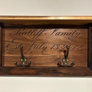 May include: A wooden wall-mounted coat rack with two hooks. The rack is made of dark wood and has a shelf on top. The rack is engraved with the text "Ratliff Family Est. July 13, 2012".