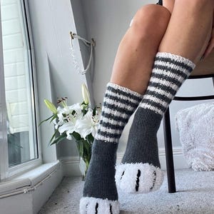 May include: Knee-high grey socks with white and grey striped cuffs and fluffy white paw-shaped bottoms. The socks are made of a soft material. The socks are worn indoors.