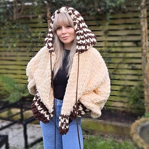 May include: A cream-coloured, oversized crochet jacket with a hood, featuring brown and white patterned accents. The jacket has a thick, textured yarn construction, with brown drawstrings. The cuffs and hood have a geometric design, offering a cosy and stylish look.