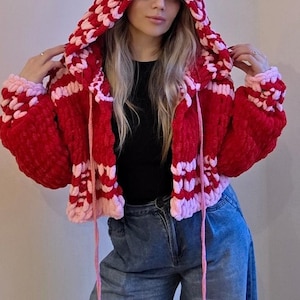 May include: A red and pink crocheted hooded jacket. The jacket has a chunky knit design with a red base and pink accents on the hood, cuffs, and trim. Pink drawstrings with tassels hang from the hood. A cosy, handmade garment.
