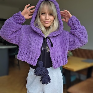 May include: A cropped, hooded jacket in a vibrant purple hue. The jacket features a chunky knit texture and a drawstring closure. Dark blue tassels hang from the front. The sleeves are long and the hood is oversized.