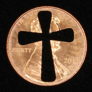 May include: A black cross cutout on a copper penny with the words "In God We Trust" and "Liberty" visible. The year "2012" is also visible on the coin.