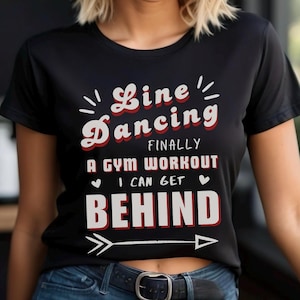 May include: Black t-shirt with white text that reads "Line Dancing Finally A Gym Workout I Can Get Behind" with a red heart on either side of the text and a black arrow pointing to the right.